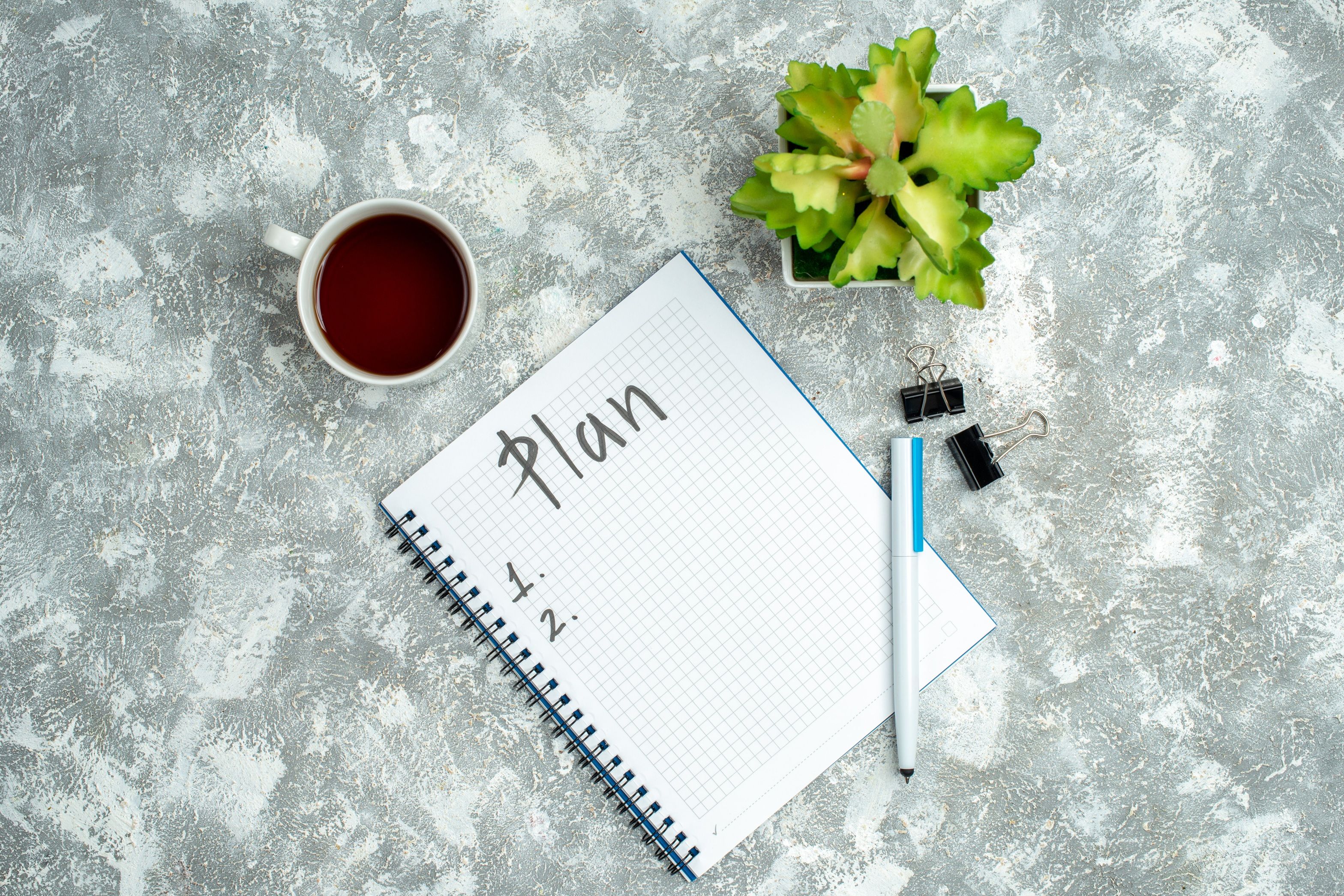 Overhead view of written spiral notebook and flower pot a cup of tea on gray background.