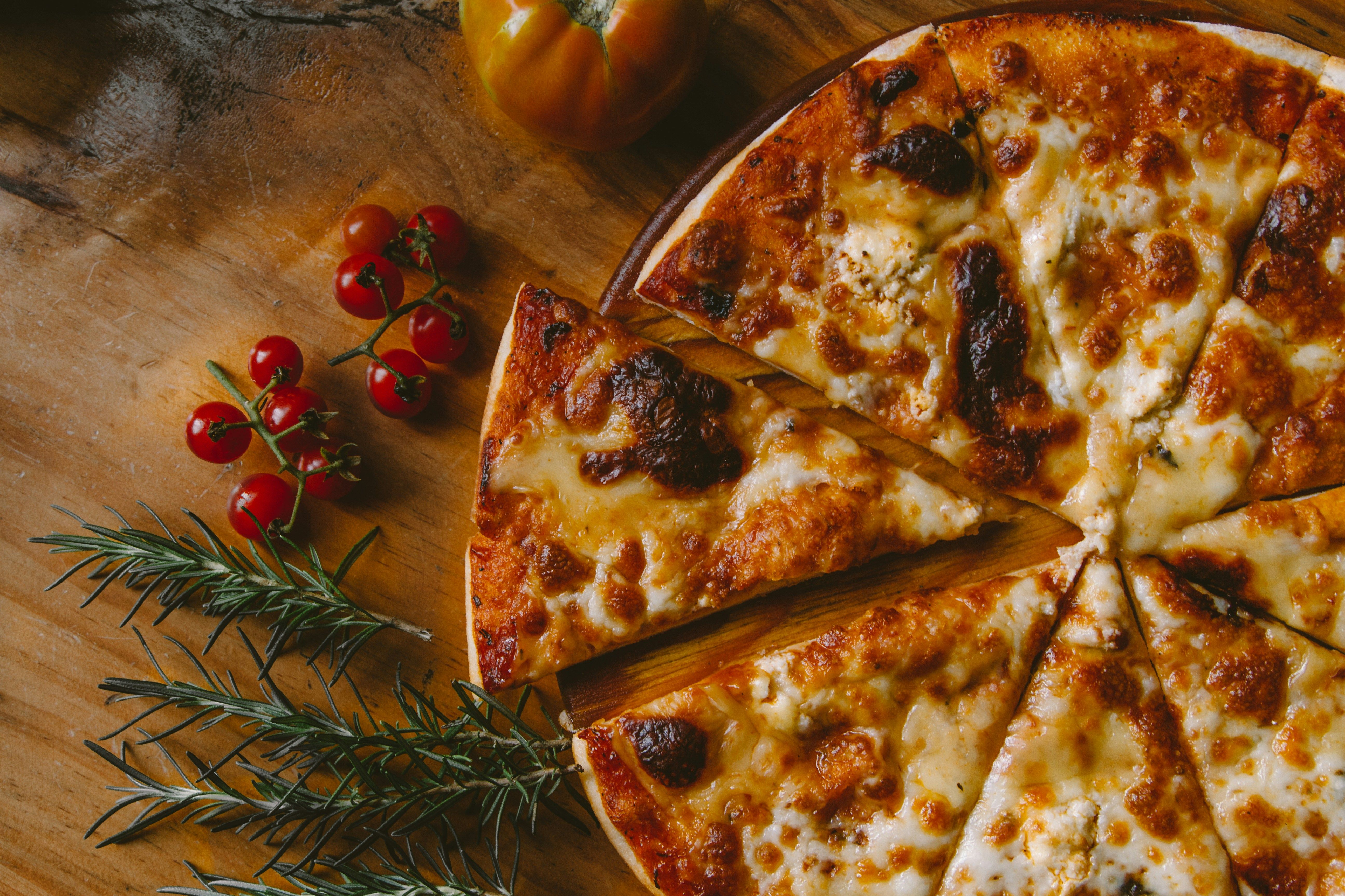 Three reosemary leaves next to some red berries, a tomato, and a pizza. They are placed on a wooden board.